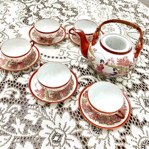 May include: A porcelain tea set featuring a teapot with a woven bamboo handle and four matching teacups with saucers. The white ceramic pieces are decorated with red and multi-colored illustrations of figures, pagodas, and floral patterns.