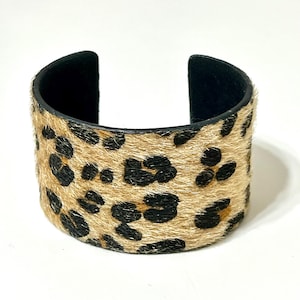 May include: A wide cuff bracelet with a leopard print design. The bracelet features a tan and black spotted pattern on the exterior and a black interior. The open design allows for easy wear.