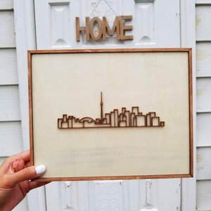 May include: A framed wood cutout of the Toronto skyline, featuring the CN Tower and other buildings. The cutout is on a light beige background and is framed in a dark wood frame.