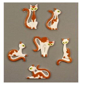 Könnte beinhalten: Sechs Keramik-Katzenfiguren in verschiedenen Posen. Die Katzen sind weiß mit braunen Flecken und haben grüne Augen.