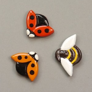 Könnte beinhalten: Drei bemalte Keramikfiguren, eine Biene, ein Marienkäfer und ein Marienkäfer mit orangenen Flügeln. Die Biene ist braun mit gelben Streifen und weißen Flügeln. Die Marienkäfer sind rot mit schwarzen Punkten und schwarzen Köpfen.