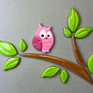 Könnte beinhalten: Keramikfliese mit einer rosa Eule auf einem braunen Ast mit grünen Blättern. Die Eule sitzt auf dem Ast mit ausgebreiteten Flügeln. Die Blätter sind um die Eule und den Ast angeordnet.
