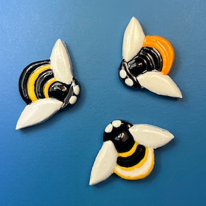 Könnte beinhalten: Drei Keramik-Hummeln mit schwarzen und gelben Streifen. Die Hummeln haben weiße Flügel und sind im Cartoon-Stil bemalt. Eine Hummel hat orangefarbene Streifen.