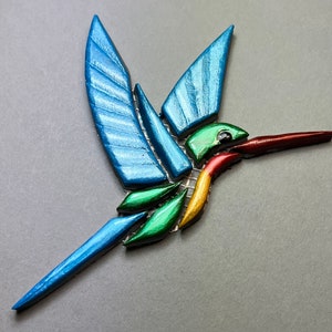 Könnte beinhalten: Eine Mosaik-Kolibri-Skulptur in Blau, Grün, Rot und Gold. Der Kolibri ist im Flug mit ausgebreiteten Flügeln.