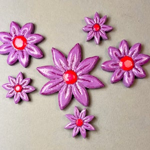 Könnte beinhalten: Sechs violette, blumenförmige Keramik-Ornamente mit roten Zentren. Die Blumen sind in Herzform auf einem grauen Hintergrund angeordnet.