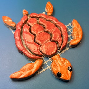 Könnte beinhalten: Eine Keramik-Mosaik-Schildkröte mit einem rosa Panzer und orangefarbenen Flossen. Die Schildkröte befindet sich auf einem weißen Netz.