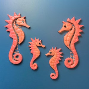 Könnte beinhalten: Vier rosafarbene Keramik-Seepferdchen-Figuren. Das größte Seepferdchen befindet sich links, das kleinste rechts und die beiden mittleren Seepferdchen in der Mitte. Alle Seepferdchen haben schwarze Augen.