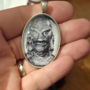 May include: Oval pendant with a silver-colored bail featuring a black and white image of a monster's face. The pendant has a shiny, reflective surface and is held in a person's hand.
