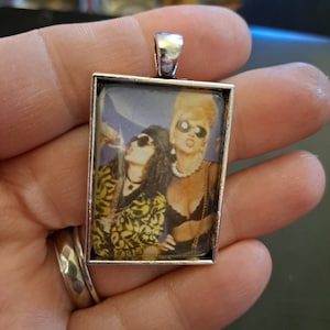 May include: A silver pendant with a glass insert featuring a photo of two women wearing sunglasses. The women are wearing colorful clothing and have their hair styled in a punk rock fashion.