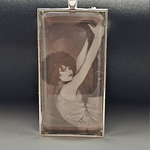 May include: Silver rectangular pendant featuring a sepia-toned image of a woman in a vintage pose. The pendant has a rectangular shape with a silver bail at the top for a chain. The woman is wearing a dress and has her arms raised.