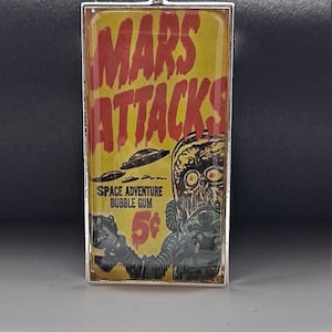 May include: A silver pendant with a glass insert featuring a vintage comic book cover for "Mars Attacks!" Space Adventure Bubble Gum. The cover features a yellow background with red text and an illustration of a Martian in a spacesuit holding a ray gun.
