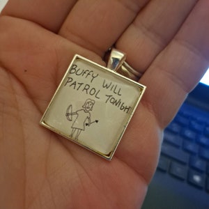 May include: Square silver pendant with a white background featuring the text "Buffy Will Patrol Tonight" and a hand-drawn illustration of a person with a bow and arrow. The pendant is held in a hand.
