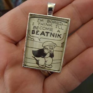 May include: Silver-toned rectangular pendant featuring a vintage comic strip image. The black and white illustration depicts a seated figure with the text "I'M BORED--I THINK I'LL BECOME A BEATNIK" in a speech bubble. The pendant is held in a hand.