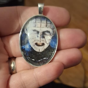 May include: A silver pendant with a round glass insert featuring a black and white image of a man's face with a stitched-up mouth. The man's face is pale and has a blue background.