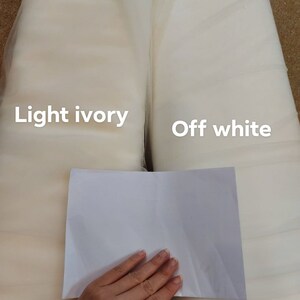 May include: Two rolls of fabric, labelled 'light ivory' and 'off white', are shown. A piece of white paper is held in front of the fabrics for colour comparison. The fabrics appear to be tulle or a similar sheer material, suitable for bridal or craft projects.