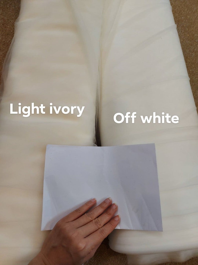 May include: Two rolls of fabric, labelled 'light ivory' and 'off white', are shown. A piece of white paper is held in front of the fabrics for colour comparison. The fabrics appear to be tulle or a similar sheer material, suitable for bridal or craft projects.