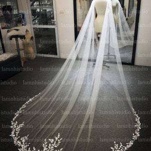 May include: A long, white bridal veil with a delicate floral lace trim. The veil is displayed on a mannequin, showcasing its flowing design. The lace trim features intricate leaf and floral patterns, adding a touch of elegance.