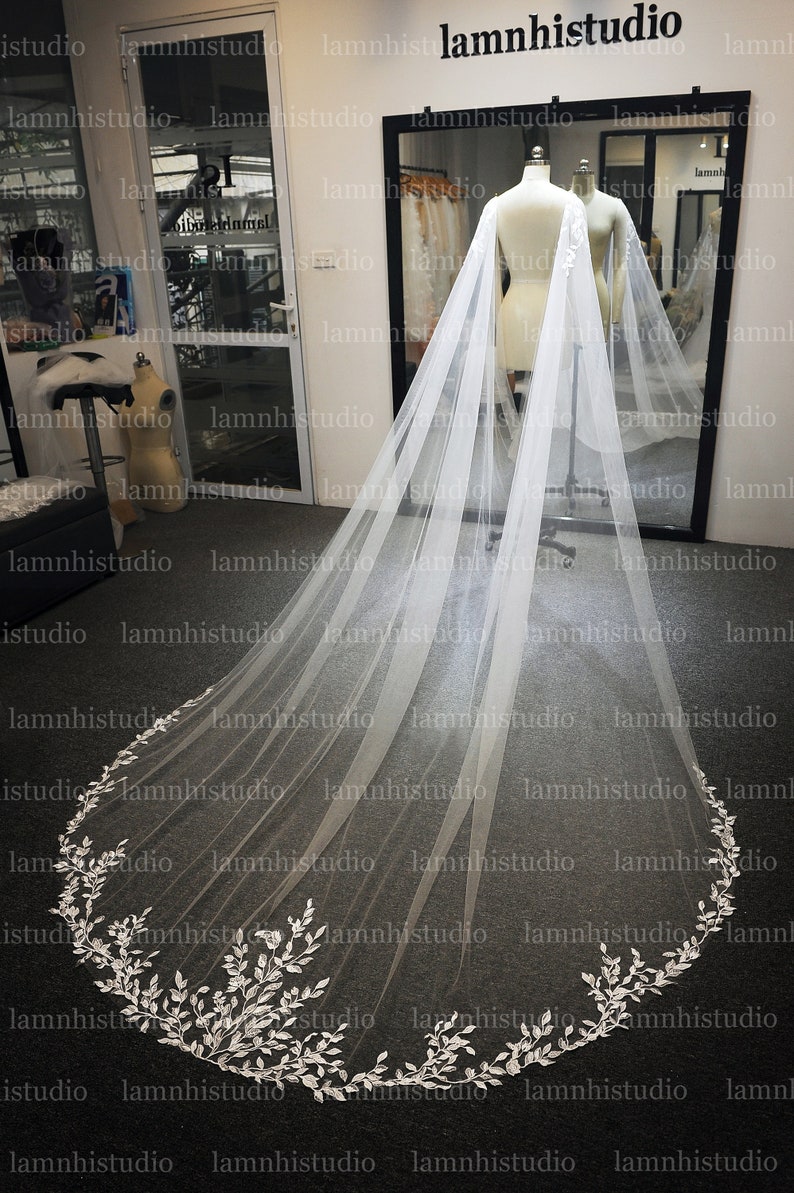May include: A long, white bridal veil with a delicate floral lace trim. The veil is displayed on a mannequin, showcasing its flowing design. The lace trim features intricate leaf and floral patterns, adding a touch of elegance.