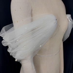 May include: White tulle sleeves adorned with faux pearls, displayed on a mannequin. The sleeves are gathered at the shoulder and wrist, creating a voluminous, layered effect. The sheer fabric allows the mannequin's form to be partially visible.