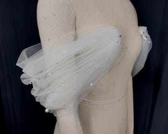 May include: White tulle sleeves adorned with faux pearls, displayed on a mannequin. The sleeves are gathered at the shoulder and wrist, creating a voluminous, layered effect. The sheer fabric allows the mannequin's form to be partially visible.