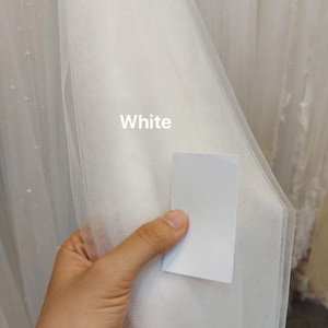 May include: A hand holds a sample of white tulle fabric, with the word "White" printed on it. The fabric is semi-transparent and has a fine mesh structure. More white tulle is visible in the background.