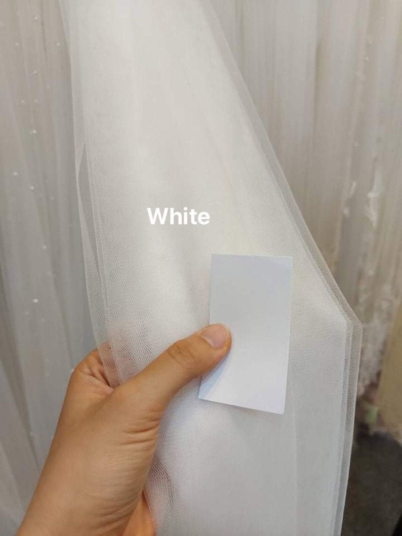 May include: A hand holds a sample of white tulle fabric, with the word "White" printed on it. The fabric is semi-transparent and has a fine mesh structure. More white tulle is visible in the background.