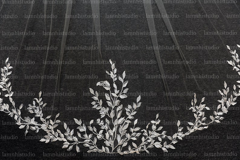 May include: A sheer white bridal veil with a delicate floral leaf design along the edge. The intricate embroidery features detailed leaves and vines, creating an elegant and romantic look. The veil is against a dark background.