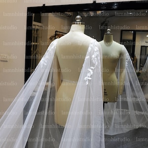 May include: A sheer white bridal cape with delicate floral lace detailing along the edges. The cape is draped over a mannequin, showcasing its flowing design. The background includes a mirror and the studio's logo.