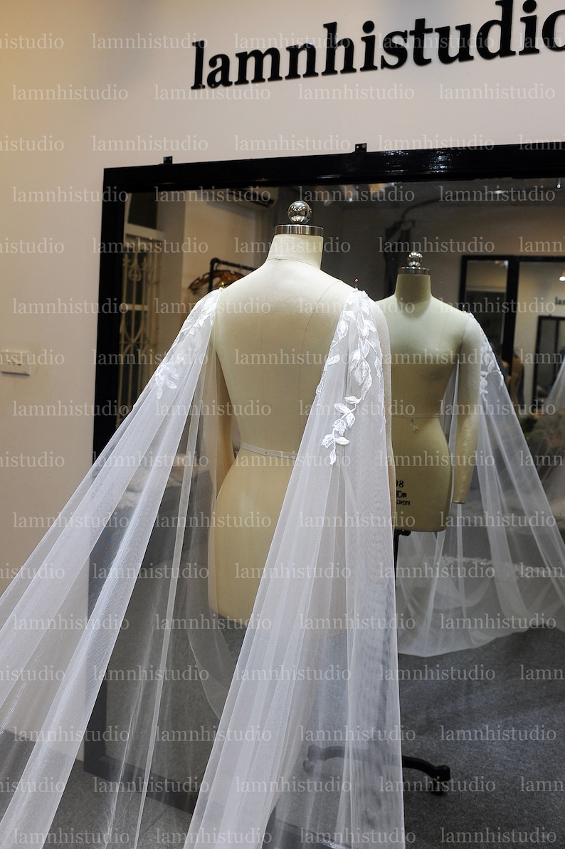May include: A sheer white bridal cape with delicate floral lace detailing along the edges. The cape is draped over a mannequin, showcasing its flowing design. The background includes a mirror and the studio's logo.
