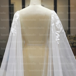 May include: A white bridal cape with delicate floral lace detailing on the shoulders. The sheer fabric drapes gracefully, creating a flowing silhouette. The cape is displayed on a mannequin, showcasing its design.