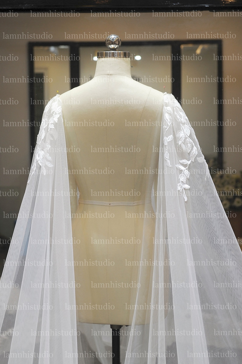 May include: A white bridal cape with delicate floral lace detailing on the shoulders. The sheer fabric drapes gracefully, creating a flowing silhouette. The cape is displayed on a mannequin, showcasing its design.