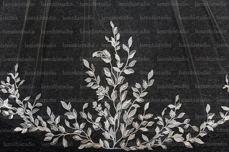 May include: A wedding veil with a detailed floral and leaf lace design. The white lace features intricate leaf shapes and a central floral element. The veil is sheer, with the lace forming a decorative border.