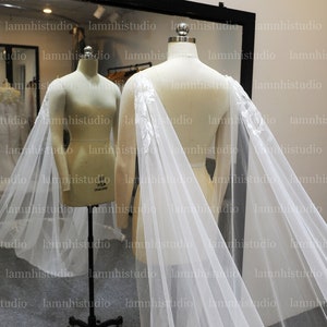 May include: A white bridal cape with floral embroidery on the shoulders is draped over a mannequin. The cape is made of a sheer, flowing fabric. The background includes a mirror and the text "lamnhistudio".