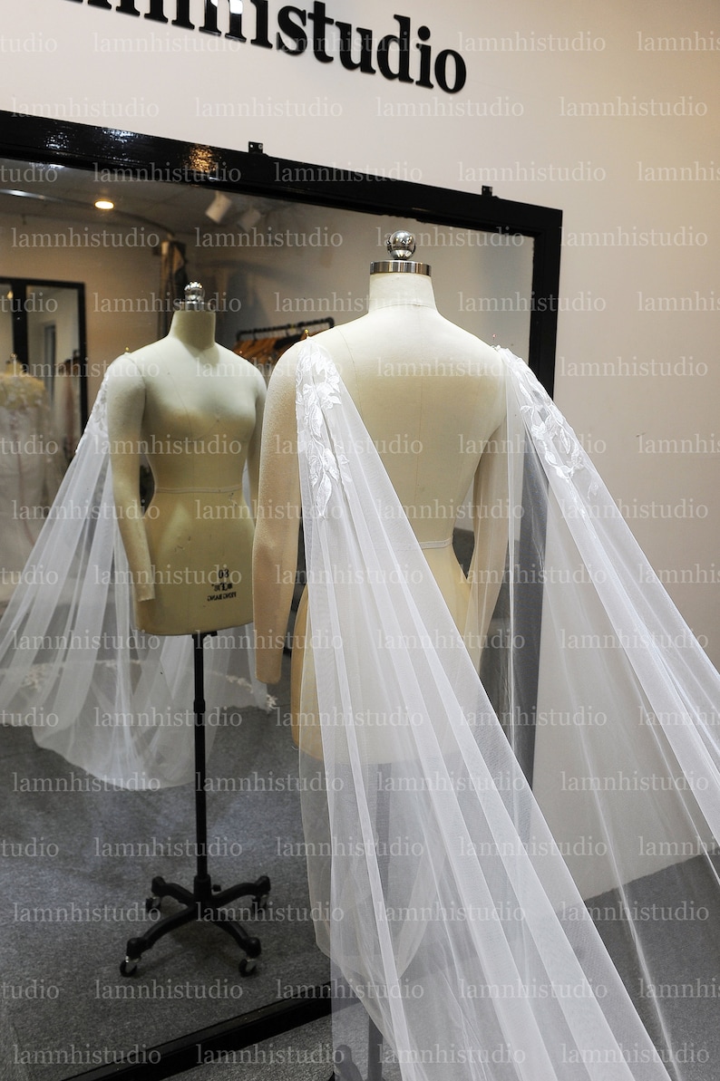 May include: A white bridal cape with floral embroidery on the shoulders is draped over a mannequin. The cape is made of a sheer, flowing fabric. The background includes a mirror and the text "lamnhistudio".