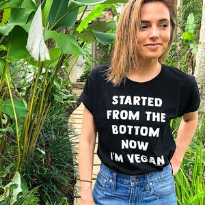 May include: A woman wearing a black t-shirt with the text "Started from the bottom now I'm vegan" in white lettering. She is standing in a garden setting with lush green foliage.