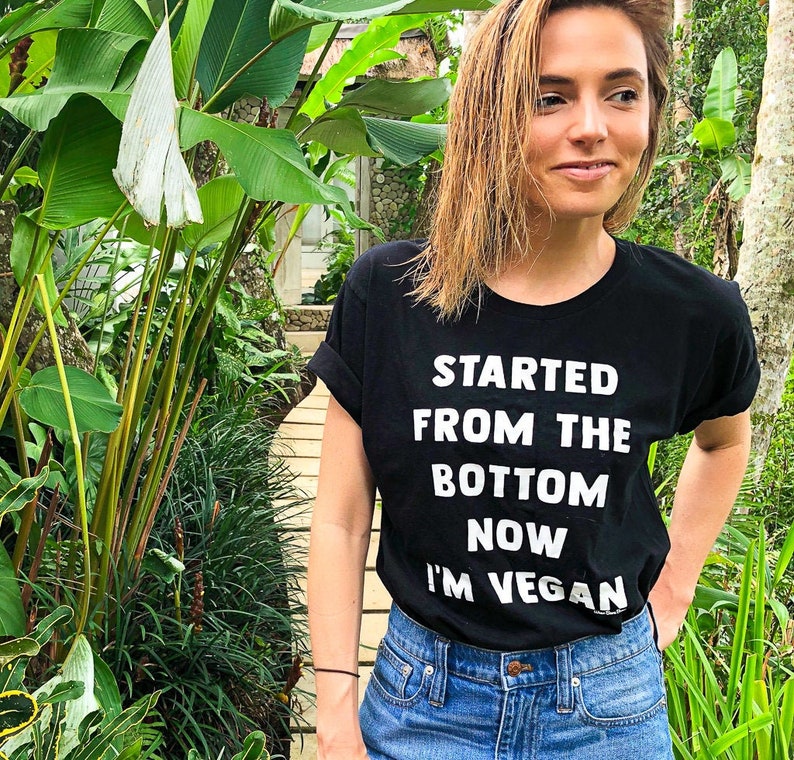 May include: A woman wearing a black t-shirt with the text "Started from the bottom now I'm vegan" in white lettering. She is standing in a garden setting with lush green foliage.