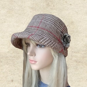 Pode incluir: Um chap&eacute;u de xadrez marrom e bege com uma borda vermelha e um pequeno pompom de pele marrom de um lado.