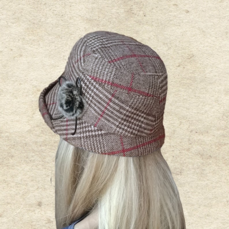 Pode incluir: Um chap&eacute;u de xadrez marrom e vermelho com um pequeno pompom de pele marrom no lado.