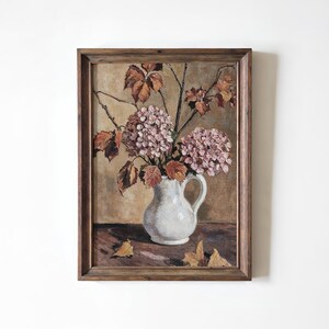 Op de afbeelding: Een ingelijst schilderij van een stilleven met een witte kan gevuld met roze hortensia's en herfstbladeren. Het kunstwerk heeft een warm kleurenpalet met bruine en beige tinten, en de lijst is van donker hout.
