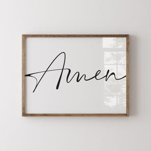 Può includere: Stampa incorniciata con la parola "Amen" in elegante scrittura nera. L'opera d'arte è ambientata su uno sfondo bianco e racchiusa in una cornice in legno naturale, adatta all'arredamento della casa.