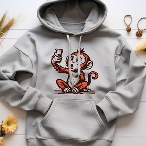 May include: A gray hoodie with a pocket on the front. The hoodie features a graphic of a monkey taking a selfie with a smartphone.