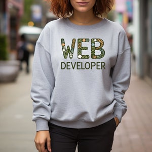 Puede incluir: Una sudadera gris con el texto "WEB DEVELOPER" en letras verdes con diseños florales. Las letras están delineadas en negro.
