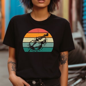 Puede incluir: Una camiseta negra con un gráfico de estilo vintage de un violonchelo frente a una puesta de sol. El gráfico está en tonos de naranja, amarillo, verde y azul.