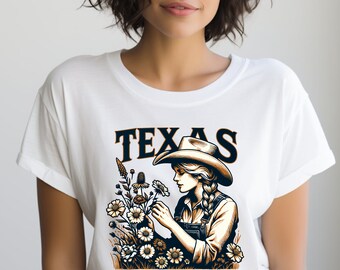 Puede incluir: Camiseta blanca con un diseño gráfico en negro y naranja que representa a una mujer con un sombrero de vaquero sosteniendo flores. El texto "TEXAS" está impreso en negro sobre el gráfico.