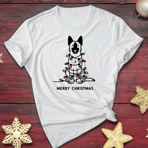 Può includere: Maglietta bianca con un'illustrazione in bianco e nero di un cane avvolto in luci di Natale. Il testo "Merry Christmas" è stampato sotto l'illustrazione.