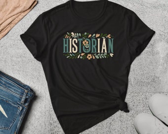 May include: Black t-shirt with the word "HISTORIAN" in a decorative font, surrounded by floral and leaf accents in green, beige, and peach. The shirt is folded and tied at the bottom.