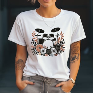 Puede incluir: Camiseta blanca con una ilustración en blanco y negro de una batería rodeada de flores marrones y blancas.
