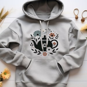 May include: A gray hoodie with a black teapot and floral design printed on the front. The teapot is surrounded by flowers and leaves in shades of blue, brown, and white.