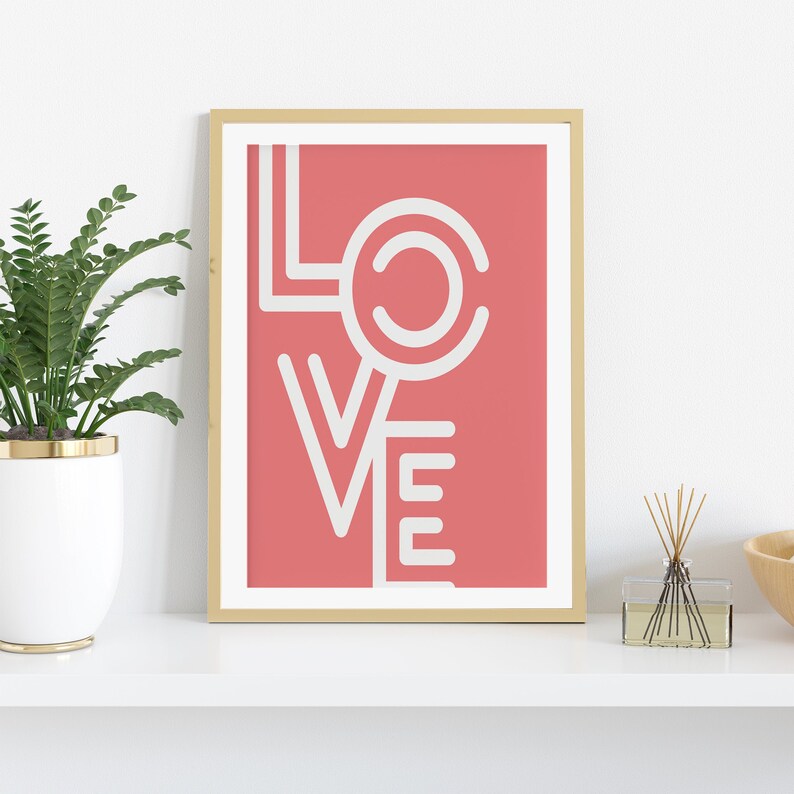 May include: A framed art print with the word "LOVE" in white, stylized block letters against a coral background. The print is in a gold frame and sits on a white shelf. A potted plant and a diffuser are also on the shelf.