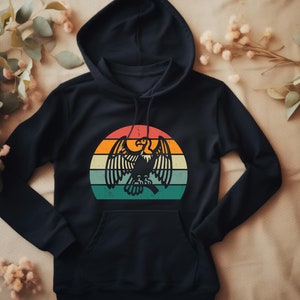 Puede incluir: Una sudadera con capucha negra con un gráfico de estilo vintage de un águila frente a una puesta de sol. El águila es negra y la puesta de sol está en tonos de naranja, amarillo y azul.